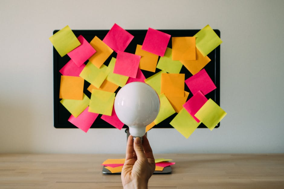 Hand holding a light bulb in front of colorful sticky notes on monitor, symbolizing brainstorming and innovation.